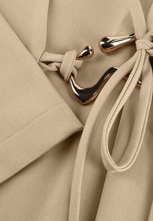 Beige stof met een gouden metalen sluiting en bijpassende beige strik, die de bevestigingsdetails van een kledingstuk benadrukt.