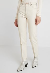 Jeans beige de talle alto con corte de pierna recta, con bolsillos delanteros, cierre de botones y costuras en contraste. Combinados con botines estampados.