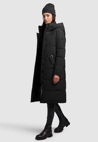Black padded coat with a hood, zip and button closure. Features a side pocket and is paired with black pants and ankle boots.