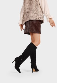 Stivali al ginocchio in suede nero con punta affusolata e tacco a stiletto. Presentano un piccolo dettaglio con fibbia e una superficie liscia.