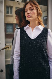 Jeune femme aux cheveux ondulés portant une chemise blanche et un gilet noir à sequins, debout devant une porte en verre réfléchissante.