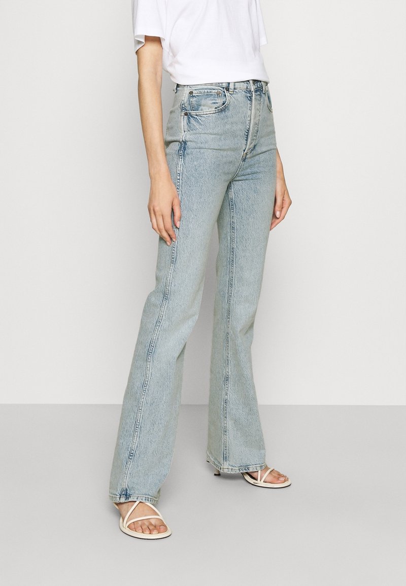 Světle modré rozšířené džíny vyrobené z denimu, s vysokým pasem a vybledlým povrchem, v kombinaci s bílým tričkem a sandály na řemínky.