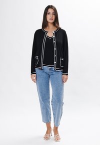 Cárdigan negro con ribete blanco, con un cuello redondo, mangas largas, dos bolsillos frontales y cierre de botones sobre una camiseta negra y unos vaqueros azules.