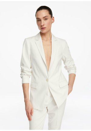 Kobieta z zaczesanymi do tyłu włosami, ubrana w rozpiętą białą marynarkę i pasujące spodnie, jedną ręką w kieszeni, na jednolitym tle.