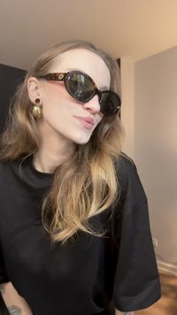 Prevelika črna sončna očala z okvirjem iz želvovega vzorca, temnimi lečami in okroglo obliko; zlati uhani v obliki obročev; razmršeni, valoviti blond lasje.