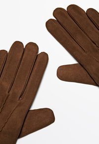 Bruine suède handschoenen tentoon gesteld met ongelijke vingers. Ze hebben een gladde textuur en decoratieve stiksels langs het polsgebied.