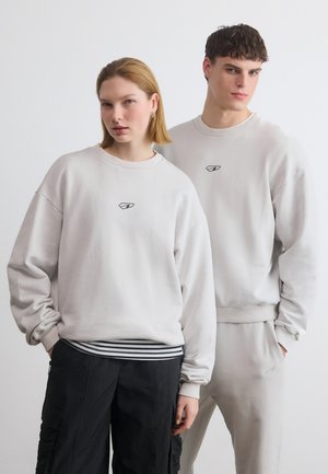 Mladá žena a muž oblečení v svetlých sivých mikinách s malými logami, stojaci blízko seba na jednoduchom bielom pozadí.