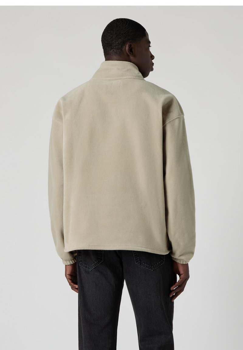 Beige fleecejacka med hög krage, nedsänkt axel och avslappnad passform, kombinerad med mörka jeans. Texturerat och mjukt material.