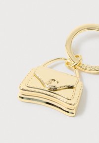 Nøglering i guldfarvet metal med en miniature håndtaskemedaljon med syninger og en lille logo-lukning på en ensfarvet baggrund.