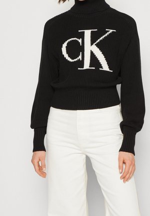 Personne portant un pull à col roulé côtelé noir avec un grand logo blanc "cK" sur le devant et un pantalon blanc taille haute.