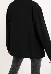 Person i en löst sittande svart långärmad tröja och urtvättade svarta skinny jeans, sedd bakifrån mot en vanlig ljus bakgrund.