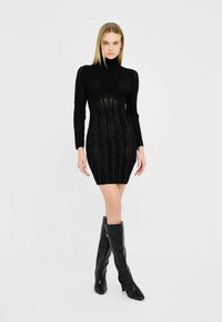 Robe en tricot noire avec des manches longues, col montant, et silhouette ajustée. Présente un motif texturé et une coupe à hauteur de cuisse. portée avec des bottes noires jusqu'aux genoux.