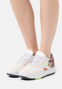 Sneakers med en blekrosa mocka ovandel, ljusgrå snören, orange och gröna accenter samt en tjock vit sula med gröna detaljer.