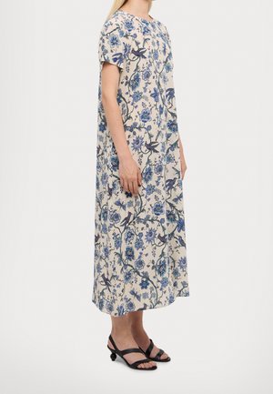 Mujer vistiendo un vestido crema de manga corta y largo hasta el tobillo con patrones florales y de pájaros en azul, combinado con sandalias negras de tacón.