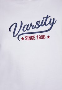 Witte t-shirt met marineblauwe cursieve borduursel van "Varsity" en rode tekst "SINCE 1998" met daaronder twee rode sterren. Gladde stoftextuur.
