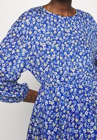 Kvinna i en blå blommig klänning med långa ärmar och en rynkad midja, som visar en tatuering på insidan av underarmen.