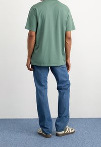 Maglietta di cotone verde a maniche corte, abbinata a jeans blu e sneakers a righe verdi, vista da dietro su uno sfondo neutro.