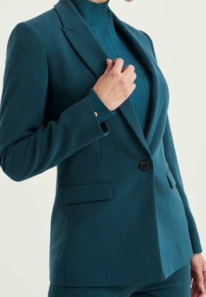 Petrolblauwe blazer met één knoop, klepzakken, gedragen over een geribde coltrui; hand die de revers van de blazer aanpast.