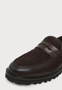 Mocassin en cuir et daim marron foncé avec une bride penny et une semelle épaisse en caoutchouc noir, vu de face.