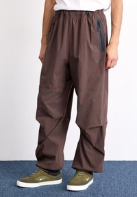 Bruna, lös passform byxor med elastisk midja och zip-ficka, tillverkade av lätt material. Kombinerat med olivgröna sneakers.