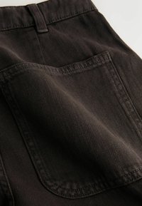 Gros plan d'une poche arrière de pantalon en denim noir avec couture visible et passant de ceinture au-dessus sur un tissu texturé.