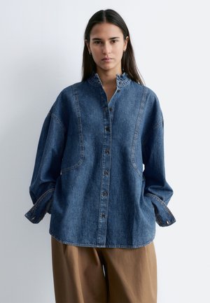 Młoda kobieta zwrócona przodem, ubrana w oversize'ową niebieską dżinsową koszulę na guziki oraz luźne brązowe spodnie, na tle jednolitego jasnego tła.