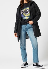 Svart vattentålig jacka över en färgglad Coca-Cola-tröja, tillsammans med ljusblå jeans och svarta högstövlade sneakers.