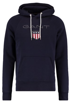 Sudadera con capucha azul marino con cordones blancos, bolsillo frontal tipo canguro, texto "GANT" y logo de escudo con estrellas y franjas en el pecho.
