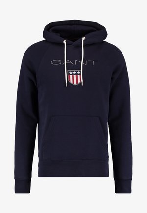 Sudadera con capucha azul marino con cordones blancos, bolsillo frontal tipo canguro, texto "GANT" y logo de escudo con estrellas y franjas en el pecho.