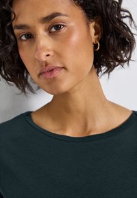 Donkergroen, kortearmige top met een ronde halslijn. De huidtextuur is glad, met subtiele make-up en kleine gouden ringoorbellen.