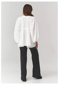 Biała bluzka z długimi rękawami i marszczonym tyłem oraz luźnym krojem, w połączeniu z czarnymi spodniami z szerokimi nogawkami na neutralnym tle.