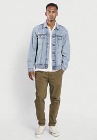 Chaqueta de mezclilla azul claro con bolsillos frontales, cierres de botón y una textura ligeramente desvaída, combinada con pantalones verde oliva y zapatillas blancas.