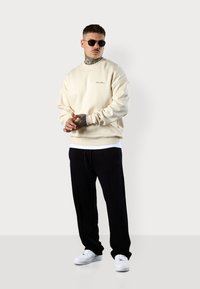 Sweatshirt crème avec des poignets et un ourlet côtelés, superposé sur une chemise blanche. Associé à un jogging noir et des baskets blanches, porté avec des lunettes de soleil.