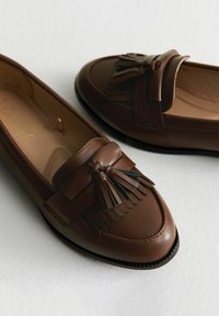 Mocasines de cuero marrón con un detalle de flecos y un acento de lazo, con una textura suave y un diseño de costura simple.