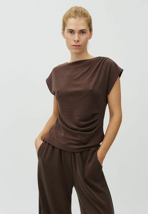 Žena s rukami v vreckách, oblečená v hnedom krátkom rukáve a voľných zodpovedajúcich nohaviciach, stojaca pred jednoduchým svetlosivým pozadím.