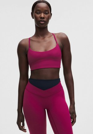 Athletische Frau trägt einen magentafarbenen Sport-BH und passende Leggings mit schwarzem Bund und steht vor einem einfarbigen hellen Hintergrund.