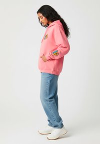 Sudadera rosa con diseños de sol bordados en amarillo y azul, con un bolsillo frontal y combinada con jeans azul claro y zapatillas blancas.