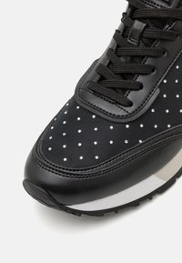 Schwarzer Sneaker mit einer strukturierten Oberseite, die kleine, runde Strasssteine enthält. Mit schwarzen Schnürsenkeln und einer weißen Mittelsohle sowie einer strukturierten Außensohle.