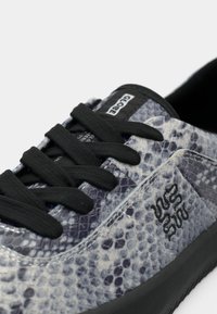 Gros plan sur une basket avec des lacets noirs, une texture en peau de serpent grise et noire, une semelle noire et une étiquette "Globe" sur la languette.