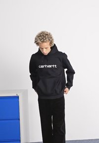 Svart hettegenser med frontlomme og justerbar hette, med en hvit "carhartt." logo på brystet, sammen med svarte bukser.