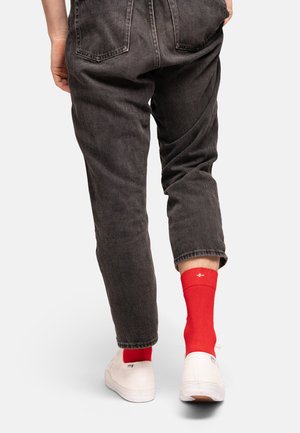 Calzini rossi con un dettaglio del logo bianco, indossati con jeans grigio scuro e scarpe di tela bianche, concentrandosi sull'area della caviglia.