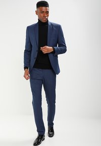 Marinblå skräddarsydd kostym med slag, slim fit, kombinerad med en svart polotröja och blanka svarta skor. Slät, texturerad tyg.