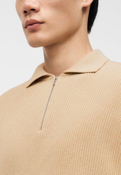 Μπεζ πλεκτό πόλο μπλουζάκι με κοντό γιακά, μπροστινό μισό φερμουάρ και υφασμάτινη υφή. Κοντινό πλάνο από τους ώμους μέχρι το πιγούνι, αναδεικνύοντας τις λεπτομέρειες.