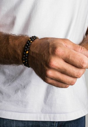Pulsera de cuentas negras y marrones con piedras de roca de lava y ojo de tigre, de ajuste elástico, llevada en una muñeca contra un fondo de camisa blanca.