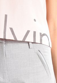 Top corto rosa chiaro con scritte nere a strisce, abbinato a pantaloni grigi con texture, dotati di tasca laterale e chiusura con zip.
