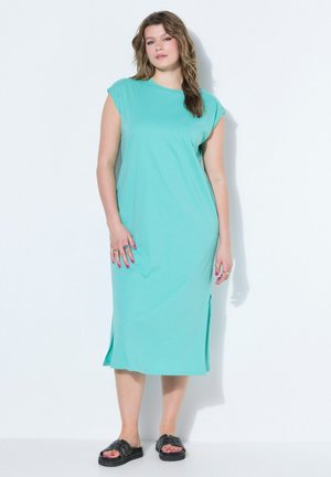 Kobieta stojąca, ubraną w turkusową sukienkę midi bez rękawów z rozcięciami po bokach oraz czarne klapki, pozująca na białym tle.