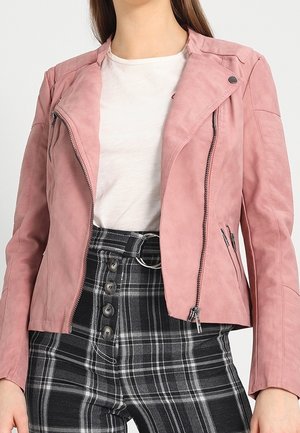 Kobieta nosząca jasnoróżową kurtkę na zamek błyskawiczny na białą koszulę, w zestawieniu z czarno-białymi spodniami w kratę z wysokim stanem z detalami w postaci guzików i paska z kółkami.