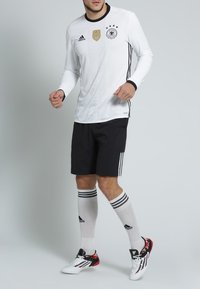 Vit fotbollströja med svarta ränder, prydd med ett guldemblem och tre stjärnor. I kombination med svarta shorts, randiga strumpor och färgglada fotbollsskor.