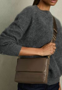 Brun læder håndtaske med flaplukning og guldfarvet hardware. Båret som crossover, parret med en grå fuzzy sweater. Simpelt og struktureret design.