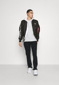 Giacca bomber nera con logo bianco sulla manica, felpa grigia sotto e pantaloni blu navy. Il modello indossa scarpe da ginnastica bianche. Design minimalista.